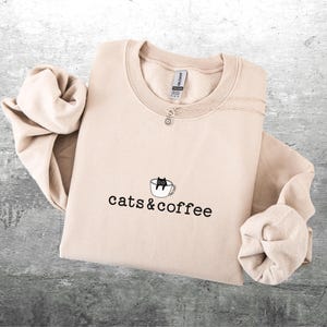 May include: A beige sweatshirt with the words "cats & coffee" printed in black. A small graphic of a black cat in a coffee cup is above the text. A silver necklace with a small charm is on the sweatshirt.