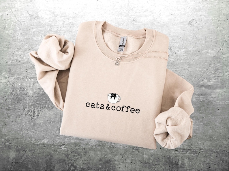 May include: A beige sweatshirt with the words "cats & coffee" printed in black. A small graphic of a black cat in a coffee cup is above the text. A silver necklace with a small charm is on the sweatshirt.