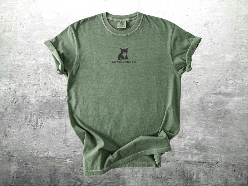May include: Olive green t-shirt with rolled sleeves. A small black cat graphic is centered on the chest, with the phrase "are you kitten me" printed below. The shirt has a vintage, worn look.