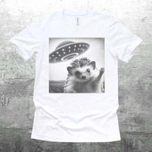 May include: A white t-shirt with a black and white image of a hedgehog waving at a flying saucer.