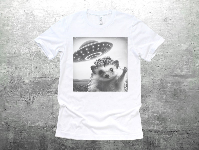 May include: A white t-shirt with a black and white image of a hedgehog waving at a flying saucer.
