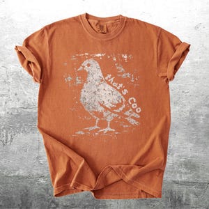 May include: An orange t-shirt with a distressed white graphic of a pigeon and the text "that's Coo". The short-sleeved shirt is made of a soft material and has rolled-up sleeves.