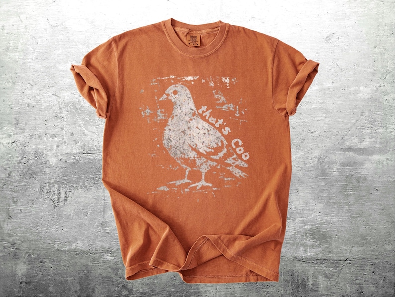 May include: An orange t-shirt with a distressed white graphic of a pigeon and the text "that's Coo". The short-sleeved shirt is made of a soft material and has rolled-up sleeves.