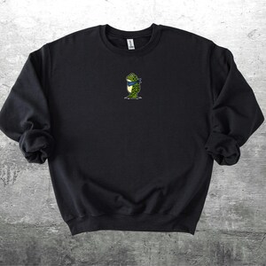 Puede incluir: Sudadera negra con un peque&ntilde;o bordado de una rana con una bufanda azul. La rana es verde con detalles amarillos. La sudadera es de cuello redondo y manga larga.