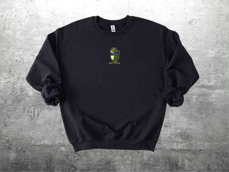 Puede incluir: Sudadera negra con un peque&ntilde;o bordado de una rana con una bufanda azul. La rana es verde con detalles amarillos. La sudadera es de cuello redondo y manga larga.