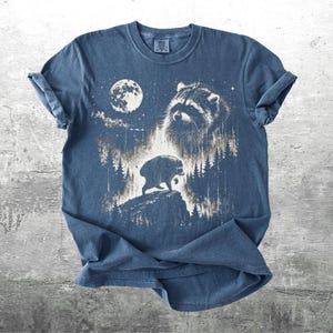 K&ouml;nnte beinhalten: Ein blaues Kurzarm-T-Shirt mit einem wei&szlig;en Grafikdruck, der ein Waschb&auml;rengesicht, einen Vollmond und die Silhouette eines Waschb&auml;ren auf einem Felsen inmitten eines Nadelwaldes zeigt. Der Text Kick Ash Co ist in der Ecke aufgedruckt.