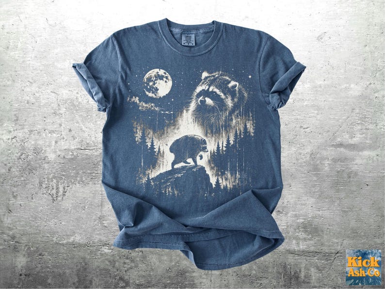 K&ouml;nnte beinhalten: Ein blaues Kurzarm-T-Shirt mit einem wei&szlig;en Grafikdruck, der ein Waschb&auml;rengesicht, einen Vollmond und die Silhouette eines Waschb&auml;ren auf einem Felsen inmitten eines Nadelwaldes zeigt. Der Text Kick Ash Co ist in der Ecke aufgedruckt.