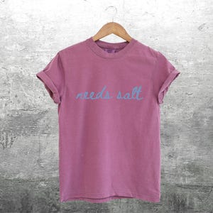 May include: A pink t-shirt with the text "needs salt" written in blue.
