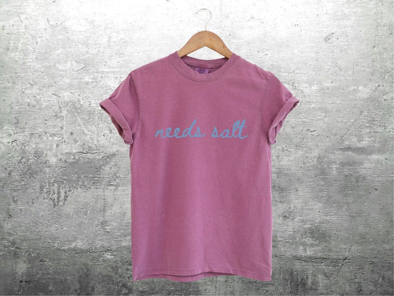 May include: A pink t-shirt with the text "needs salt" written in blue.