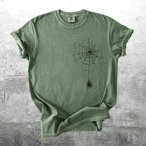 May include: Sage green t-shirt featuring a black spider web design with a spider hanging below. The short-sleeved shirt is made of a soft material and is displayed on a gray surface. The shirt has a classic crew neck.