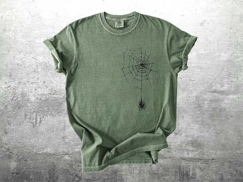 May include: Sage green t-shirt featuring a black spider web design with a spider hanging below. The short-sleeved shirt is made of a soft material and is displayed on a gray surface. The shirt has a classic crew neck.