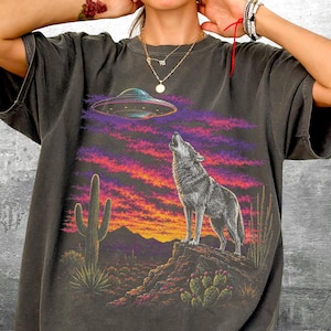 May include: A charcoal gray t-shirt featuring a graphic print of a wolf howling at a UFO in a desert landscape. The scene includes a saguaro cactus, rocky terrain, and a vibrant sunset sky with purple and orange hues. The text Kick Ash Co is visible.