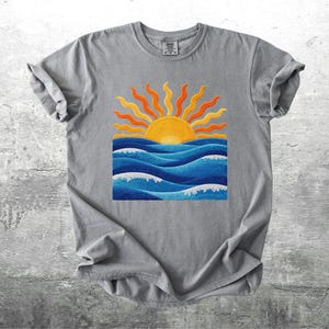 May include: A gray t-shirt featuring a vibrant graphic of a sunset over ocean waves. The sun is yellow and orange, with blue waves and white foam. The shirt is made of soft material.