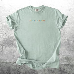 Puede incluir: Una camiseta verde claro con el texto "It's my birthday" en colores arco&iacute;ris.