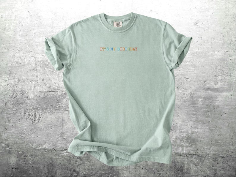 Puede incluir: Una camiseta verde claro con el texto "It's my birthday" en colores arco&iacute;ris.