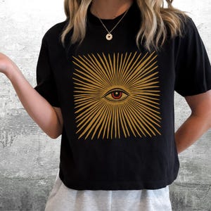 May include: A black short-sleeve t-shirt featuring a centered graphic of a stylized eye surrounded by radiating golden sunburst lines. The design is printed in a warm yellow-gold tone against the solid black fabric of the garment.