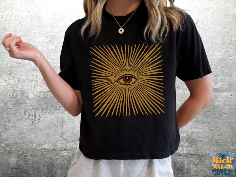 May include: A black short-sleeve t-shirt featuring a centered graphic of a stylized eye surrounded by radiating golden sunburst lines. The design is printed in a warm yellow-gold tone against the solid black fabric of the garment.