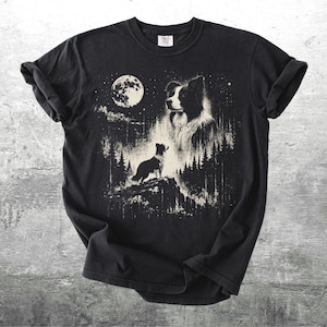 Pu&ograve; includere: T-shirt nero con una grafica dettagliata di un cane Border Collie sotto la luna piena. Il design include una silhouette di cane su una collina, alberi e stelle, creando una scena notturna.
