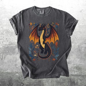 May include: A dark gray t-shirt with a black dragon illustration. The dragon has orange and yellow wings and is surrounded by floral designs in shades of blue, orange, and yellow.
