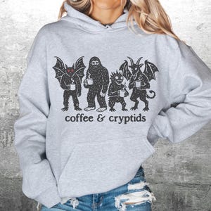 May include: A light gray hoodie with a black graphic of four cryptids holding coffee mugs. The text "coffee & cryptids" is printed below the figures. The hoodie has a front pocket and a drawstring hood.