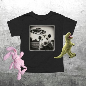 May include: Black toddler t-shirt with a sepia-toned image of an opossum under a UFO. A pink stuffed bunny and a green stuffed dinosaur are on the sides of the shirt.