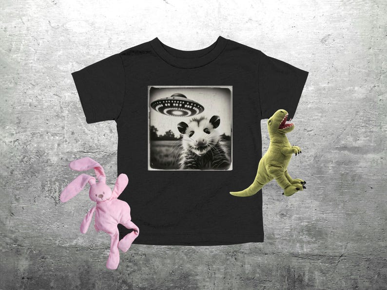 May include: Black toddler t-shirt with a sepia-toned image of an opossum under a UFO. A pink stuffed bunny and a green stuffed dinosaur are on the sides of the shirt.