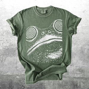 May include: A sage green t-shirt featuring a white graphic of a frog's face with spiral eyes. The shirt has rolled-up sleeves and a crew neck. The design is a bold, eye-catching illustration.
