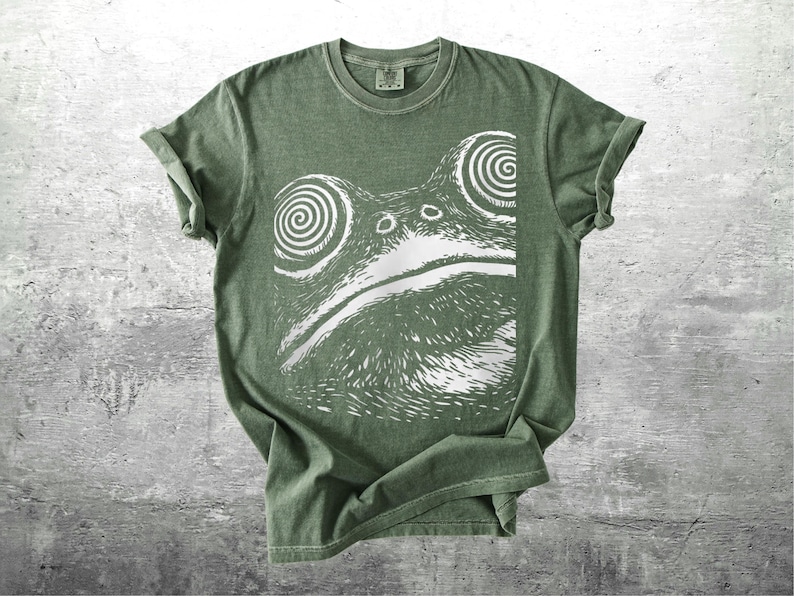 May include: A sage green t-shirt featuring a white graphic of a frog's face with spiral eyes. The shirt has rolled-up sleeves and a crew neck. The design is a bold, eye-catching illustration.