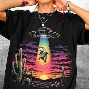 May include: A black t-shirt featuring a graphic print of a possum being abducted by a UFO. The scene includes a desert landscape with cacti under a sunset sky with purple and pink clouds. The text Kick Ash Co is printed in the bottom right corner.