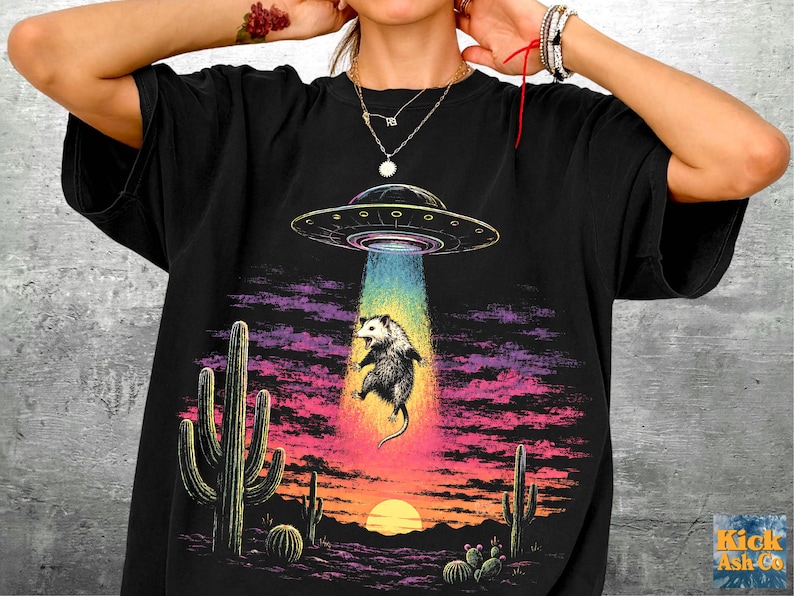 May include: A black t-shirt featuring a graphic print of a possum being abducted by a UFO. The scene includes a desert landscape with cacti under a sunset sky with purple and pink clouds. The text Kick Ash Co is printed in the bottom right corner.