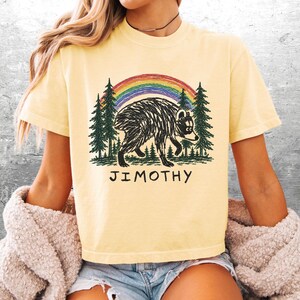 May include: A pale yellow short-sleeve t-shirt featuring a graphic print of a black bear walking through a forest of green pine trees under a colorful rainbow. The text JIMOTHY is printed in black capital letters below the bear illustration.