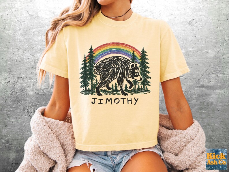 May include: A pale yellow short-sleeve t-shirt featuring a graphic print of a black bear walking through a forest of green pine trees under a colorful rainbow. The text JIMOTHY is printed in black capital letters below the bear illustration.