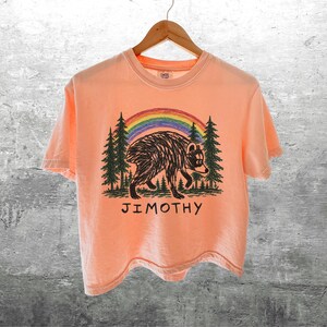 May include: A peach-colored short-sleeve t-shirt featuring a graphic of a black bear walking through a forest of green pine trees under a colorful rainbow. The name JIMOTHY is printed in black capital letters below the bear illustration.