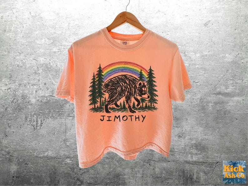 May include: A peach-colored short-sleeve t-shirt featuring a graphic of a black bear walking through a forest of green pine trees under a colorful rainbow. The name JIMOTHY is printed in black capital letters below the bear illustration.