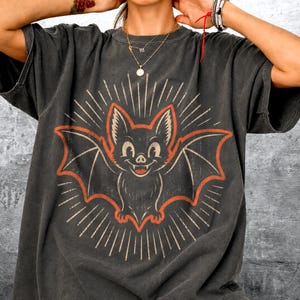May include: A charcoal gray short-sleeve t-shirt featuring a central graphic of a smiling bat with orange outlines and radiating white lines. The bat has pointed ears and visible fangs. The fabric has a faded, washed-out appearance.