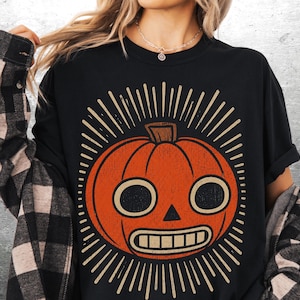 May include: A black t-shirt featuring a large, centered graphic of an orange pumpkin with a stylized face. The pumpkin has two large circular eyes, a triangular nose, and a rectangular mouth with teeth. Sunburst lines radiate outward from the pumpkin.