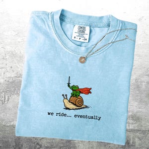 May include: Light blue t-shirt with a graphic of a frog riding a snail, holding a sword and wearing a cape. The text "we ride... eventually" is below the graphic. A silver necklace with a pendant is on top.