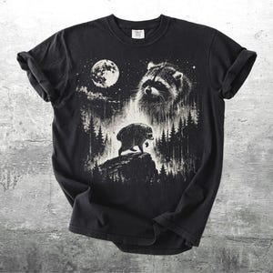 K&ouml;nnte beinhalten: Ein schwarzes Kurzarm-T-Shirt mit einem wei&szlig;en Grafikdruck eines Waschb&auml;ren, der nachts auf einer Felsklippe in einem Wald steht. Das Design zeigt einen gro&szlig;en Vollmond, ein detailliertes Waschb&auml;renportr&auml;t und Tannenb&auml;ume.