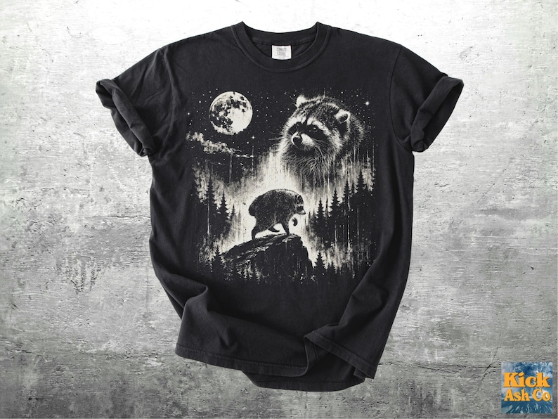 K&ouml;nnte beinhalten: Ein schwarzes Kurzarm-T-Shirt mit einem wei&szlig;en Grafikdruck eines Waschb&auml;ren, der nachts auf einer Felsklippe in einem Wald steht. Das Design zeigt einen gro&szlig;en Vollmond, ein detailliertes Waschb&auml;renportr&auml;t und Tannenb&auml;ume.