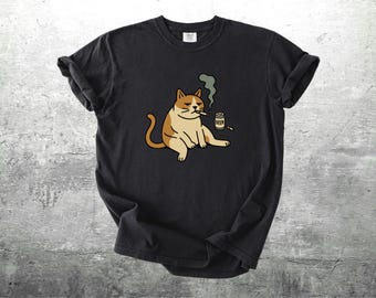 May include: Black t-shirt featuring a cartoon cat with a cigarette in its mouth, exhaling smoke. The cat is tan and white, sitting next to a small beer can. The shirt is made of a soft material.