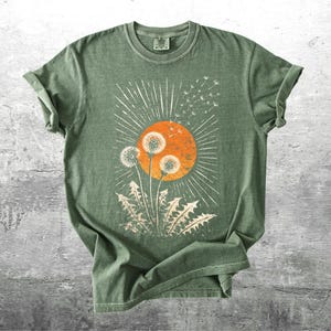 May include: A sage green t-shirt featuring a graphic of dandelions in front of an orange sun with white rays. The dandelion design is in a cream color. The shirt has rolled-up sleeves and a crew neck.
