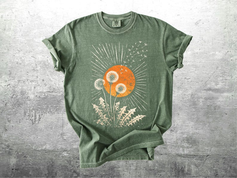 May include: A sage green t-shirt featuring a graphic of dandelions in front of an orange sun with white rays. The dandelion design is in a cream color. The shirt has rolled-up sleeves and a crew neck.