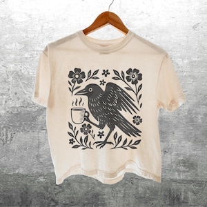 May include: Cream-colored t-shirt featuring a black graphic of a crow holding a coffee cup, surrounded by floral elements. The design is centered on the shirt. The shirt is hanging on a wooden hanger.
