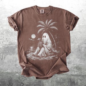 May include: A brown short-sleeve t-shirt featuring a white graphic print of a ghost sitting on a beach under a palm tree, holding a pineapple drink with a small umbrella. The scene includes a full moon over the water in the background.