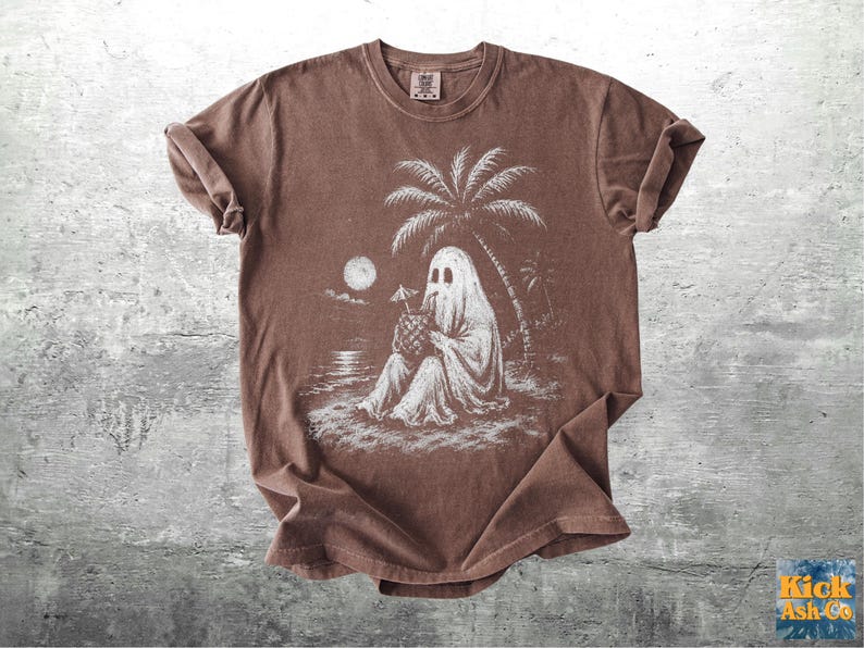 May include: A brown short-sleeve t-shirt featuring a white graphic print of a ghost sitting on a beach under a palm tree, holding a pineapple drink with a small umbrella. The scene includes a full moon over the water in the background.