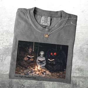 May include: Gray t-shirt with a graphic of Bigfoot, an alien, and a moth-like creature reading books around a campfire. The text "Late Night Readers Club" is printed below the image. A silver necklace with a pendant is draped on the shirt.