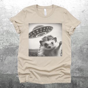 May include: A beige t-shirt with a black and white image of a hedgehog waving at a flying saucer.