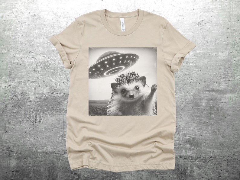 May include: A beige t-shirt with a black and white image of a hedgehog waving at a flying saucer.