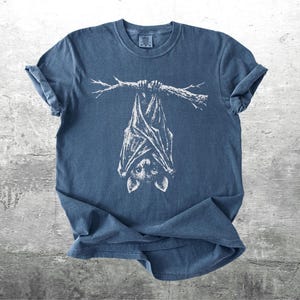 Op de afbeelding: Blauw t-shirt met een witte illustratie van een vleermuis die ondersteboven aan een tak hangt. De vleermuis heeft gedetailleerde vleugels en een schattig gezicht. Het shirt is gemaakt van zacht materiaal en heeft opgerolde mouwen.
