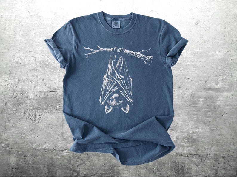 Op de afbeelding: Blauw t-shirt met een witte illustratie van een vleermuis die ondersteboven aan een tak hangt. De vleermuis heeft gedetailleerde vleugels en een schattig gezicht. Het shirt is gemaakt van zacht materiaal en heeft opgerolde mouwen.